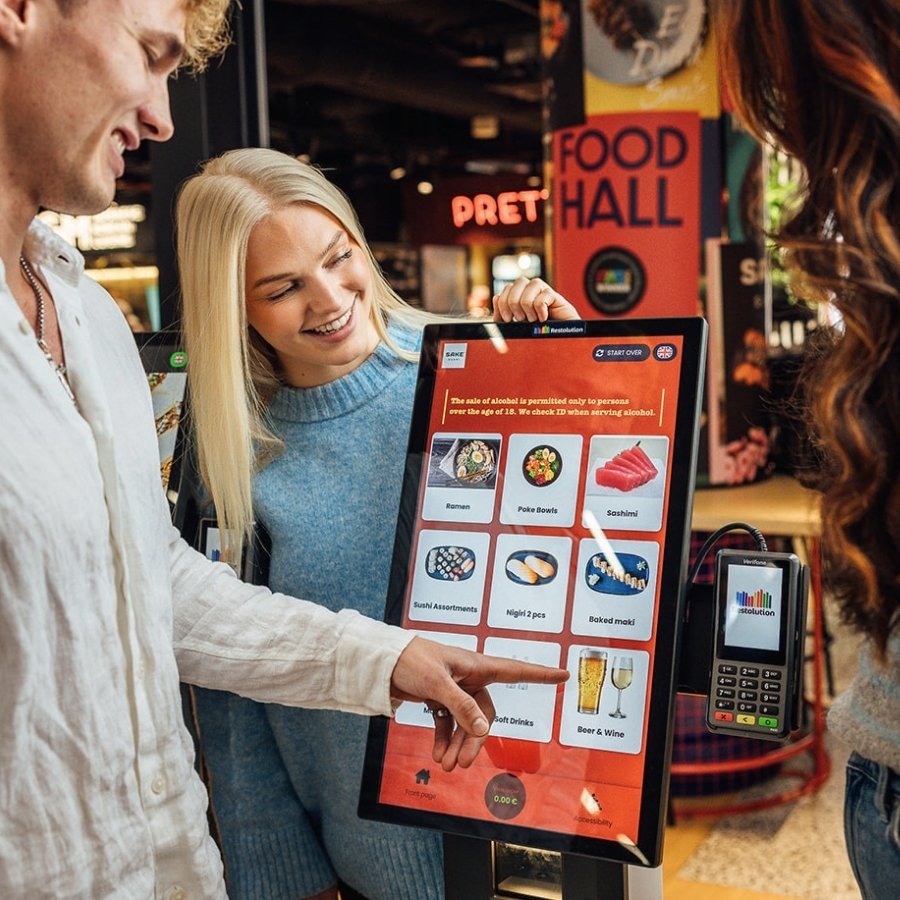 Two people using a touchscreen self-service kiosk in a food hall to order sushi and drinks.
