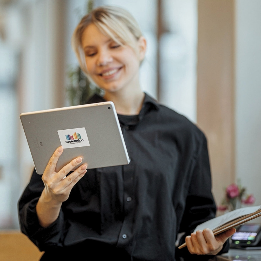 Smiling woman in black shirt holding a tablet and an open notebook indoors.