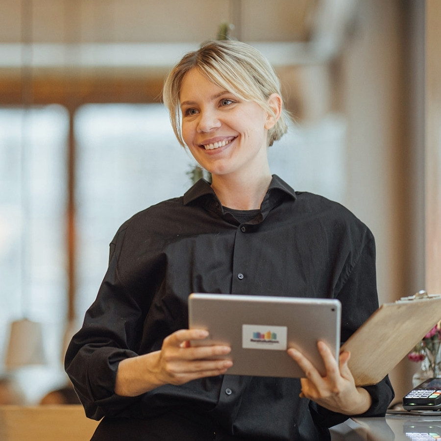 Smiling woman in black shirt holding tablet and clipboard in a bright office setting.