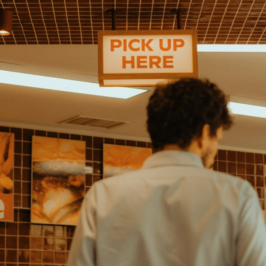 Back view of a man standing under a 'Pick Up Here' sign inside a restaurant.