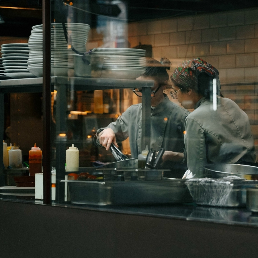 Two chefs in gray uniforms preparing food in a kitchen behind a glass window with stacked plates.