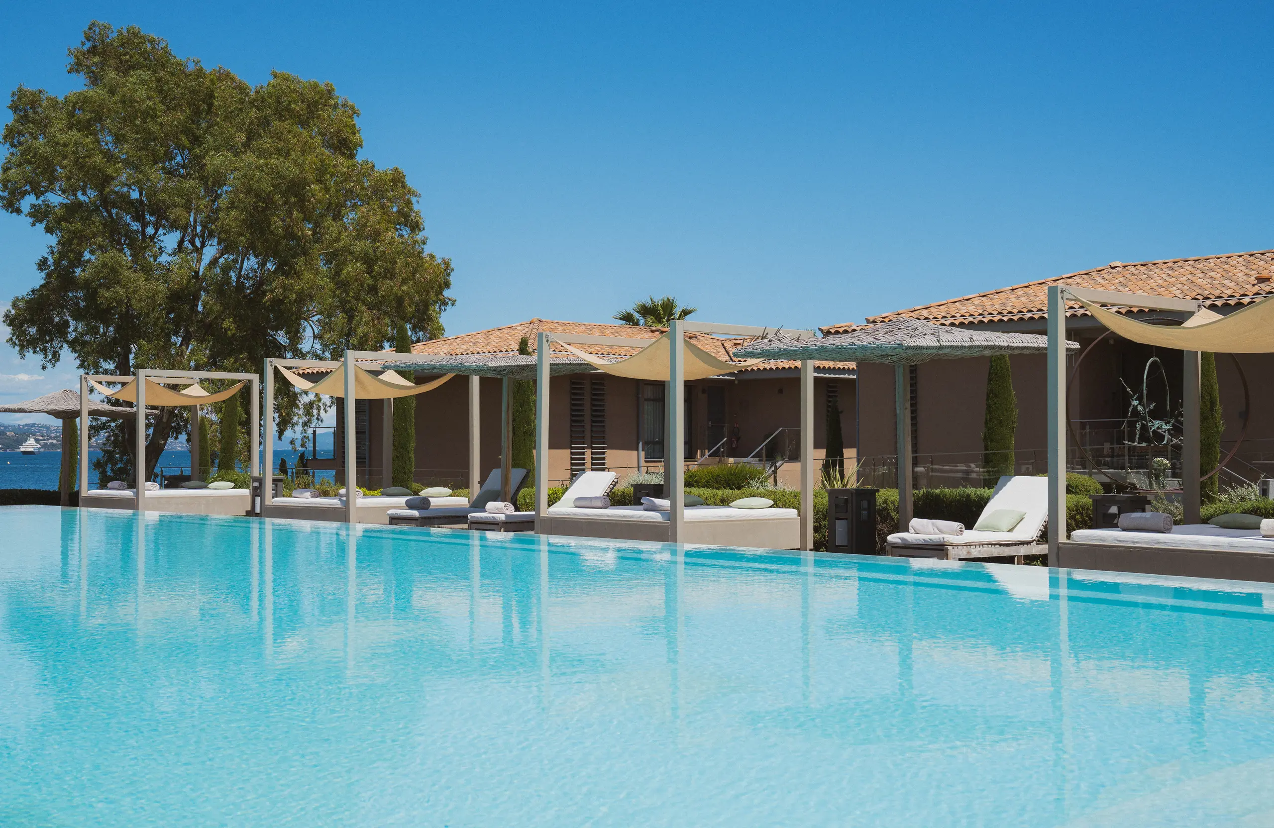 Piscine à débordement avec des chaises longues sous des pergolas près d'une maison à toit en tuiles.