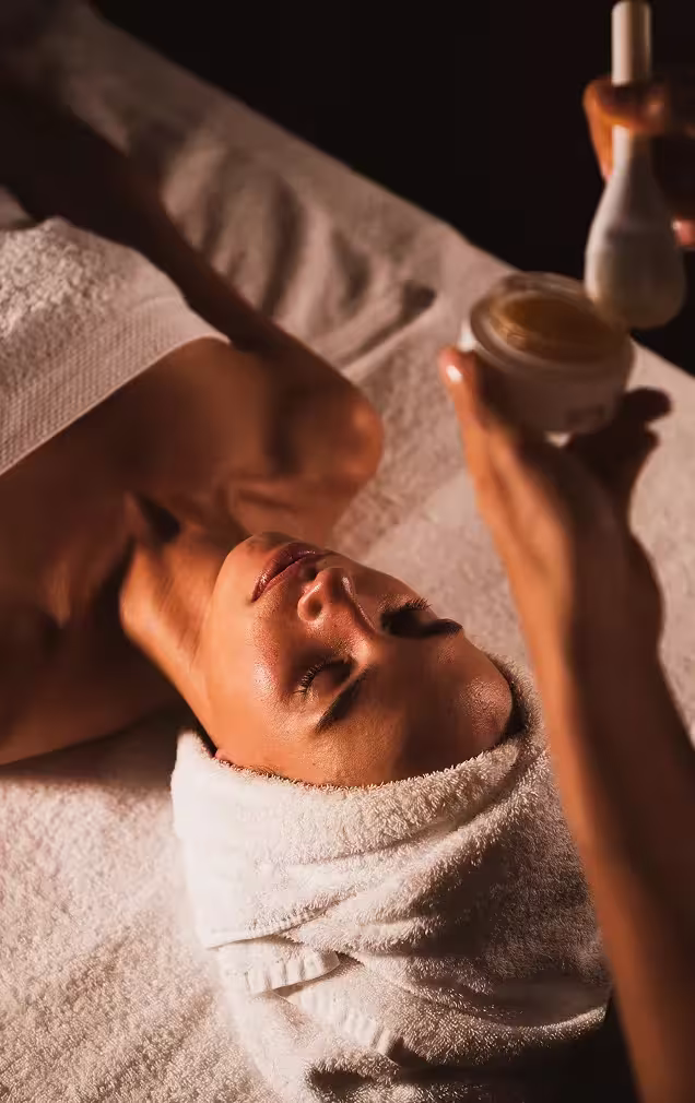 Femme recevant un soin du visage avec une brosse applicatrice dans un spa, allongée sur une serviette blanche, les yeux fermés et une serviette enroulée autour de la tête.