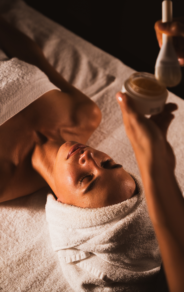 Femme recevant un soin du visage avec une brosse applicatrice dans un spa, allongée sur une serviette blanche, les yeux fermés et une serviette enroulée autour de la tête.
