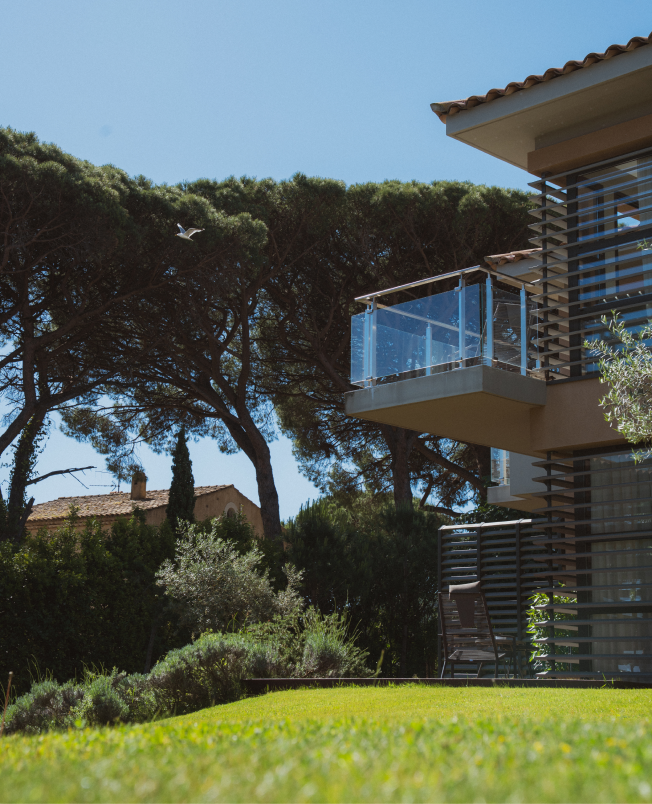 Balcon en verre d'une maison moderne avec jardin vert, arbres et ciel bleu clair en arrière-plan.