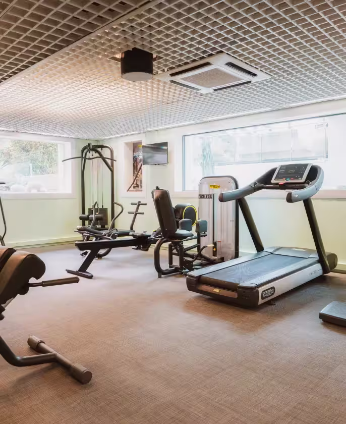 Salle de sport lumineuse avec tapis roulant, équipements de musculation et grandes fenêtres.