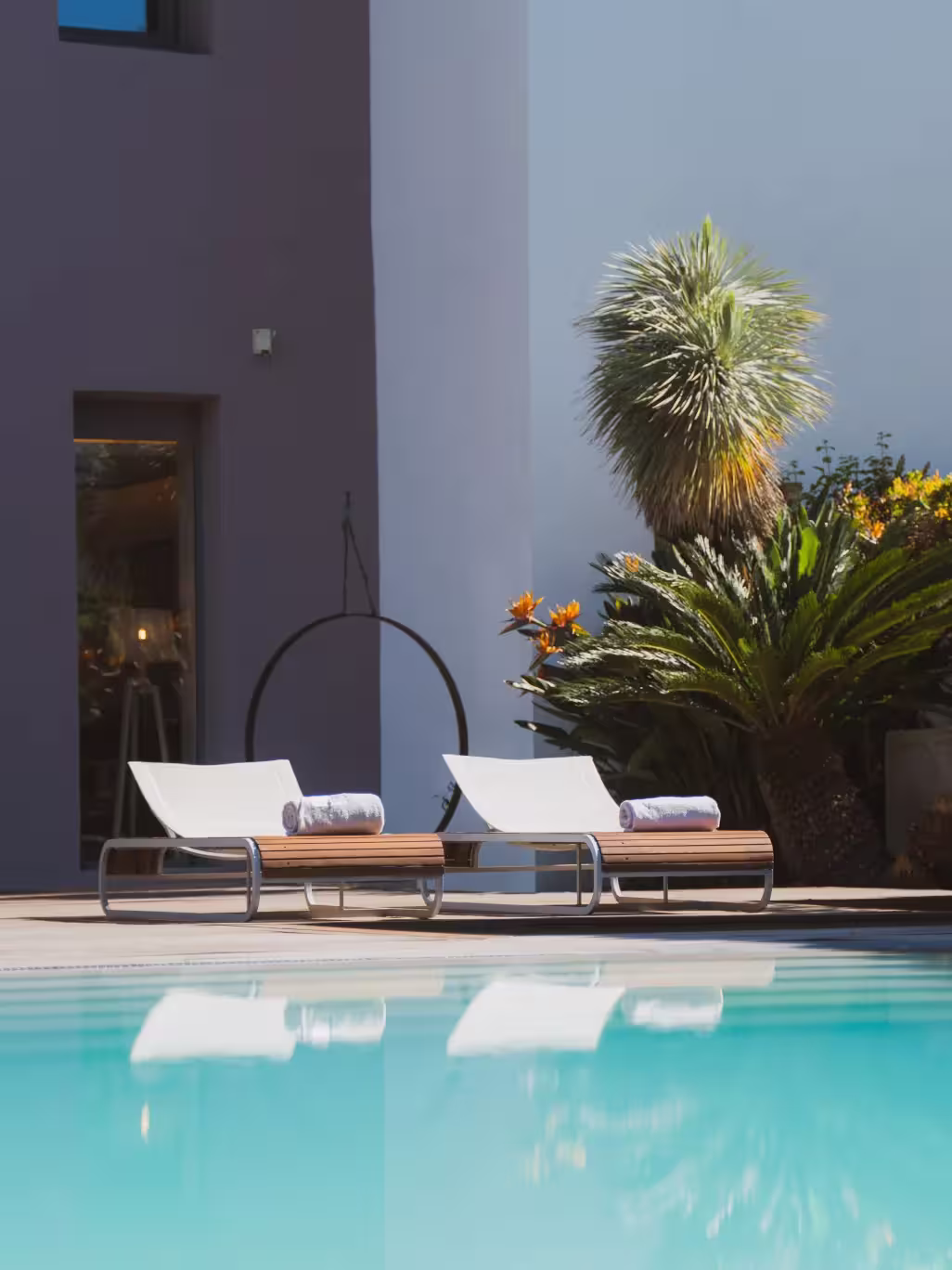 Deux chaises longues blanches avec serviettes roulées au bord d'une piscine bleue, entourées de plantes vertes et fleurs orange.