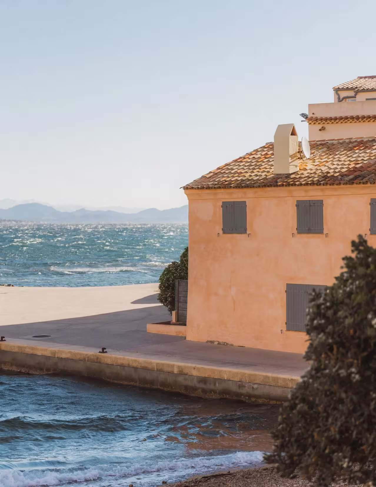 Maison en bord de mer avec toit en tuiles et volets fermés, devant une mer agitée et des montagnes en arrière-plan.