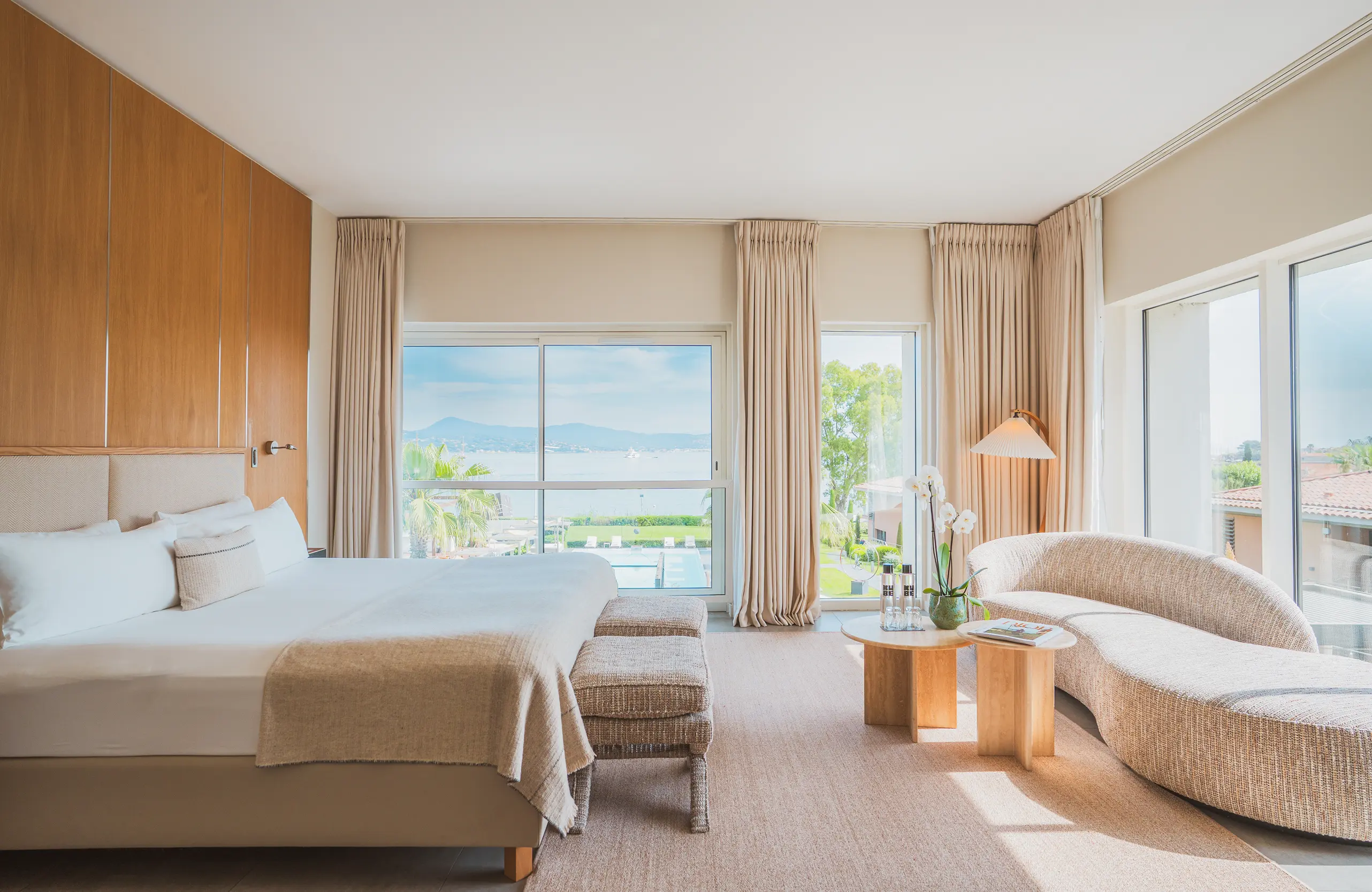 Chambre d'hôtel lumineuse avec grand lit, canapé incurvé, tables basses en bois, et vue sur la mer à travers de grandes fenêtres.