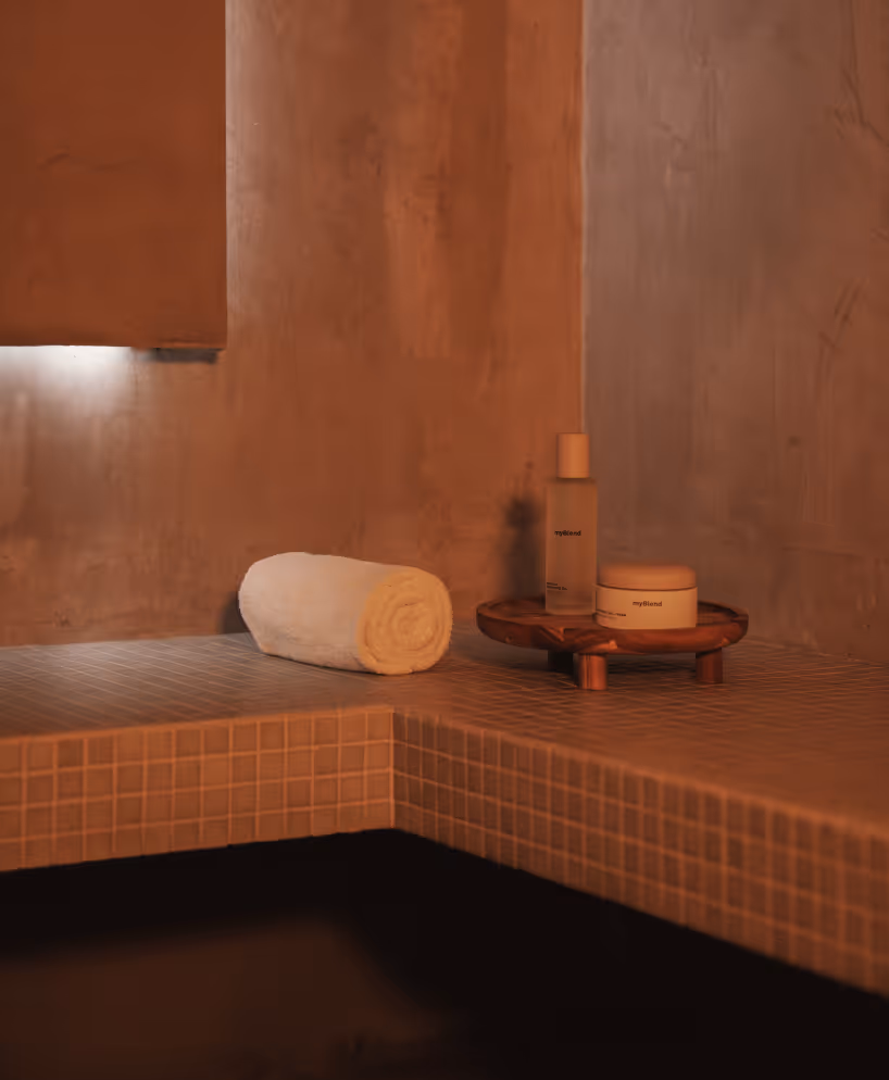 Coin spa avec un banc en carrelage beige, une serviette blanche roulée et des produits de soin myBlend sur un plateau en bois.