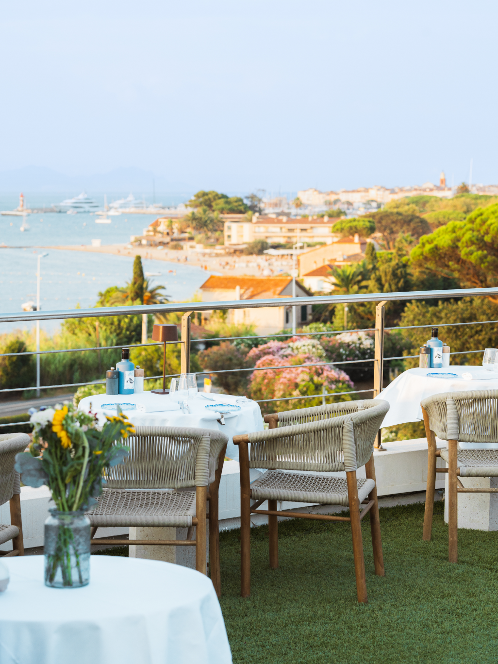 Terrasse de restaurant avec tables en nappes blanches et chaises en corde, vue sur la mer, bateaux et village en arrière-plan.