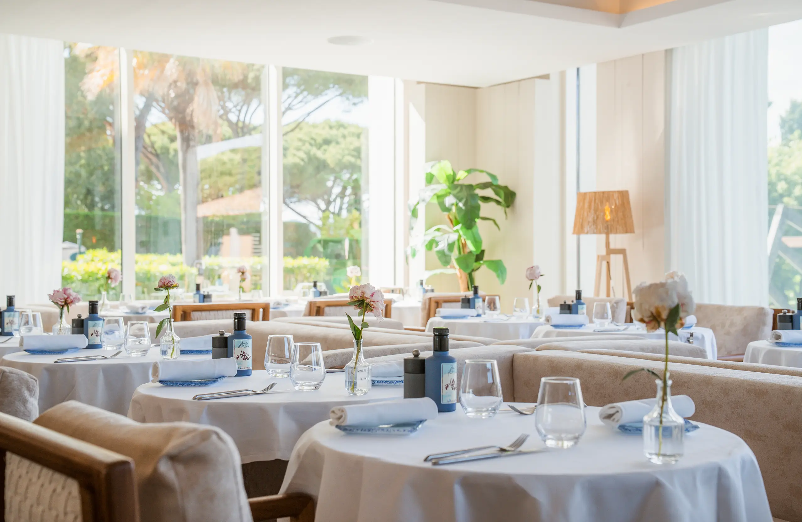 Salle de restaurant élégante avec tables rondes dressées de nappes blanches, verres, serviettes roulées et fleurs dans des petits vases, éclairée par une grande baie vitrée.