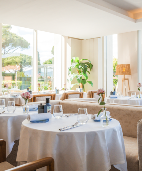 Salle de restaurant lumineuse avec grandes fenêtres, tables rondes nappées, décorées de petites fleurs roses et verres, chaises beiges et plante verte dans un coin.