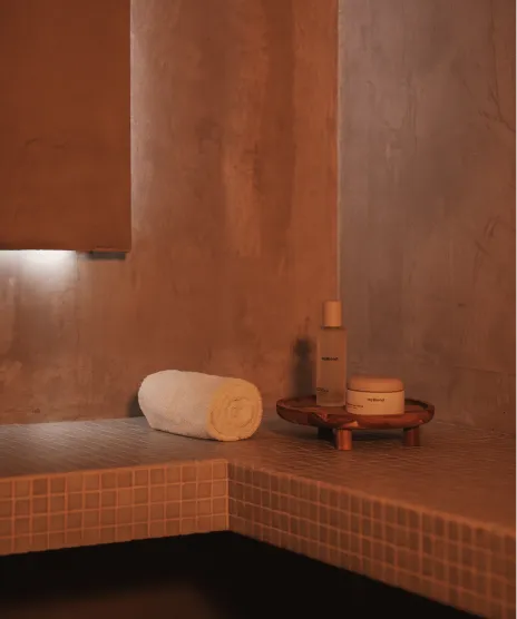 Coin d'une salle de bain avec un banc carrelé, une serviette blanche roulée et des produits de soin sur un plateau en bois.