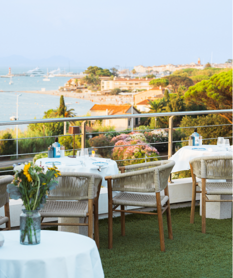 Terrasse de restaurant avec tables dressées, chaises en bois tressé, vue sur la mer, des bateaux et des bâtiments côtiers au coucher du soleil.
