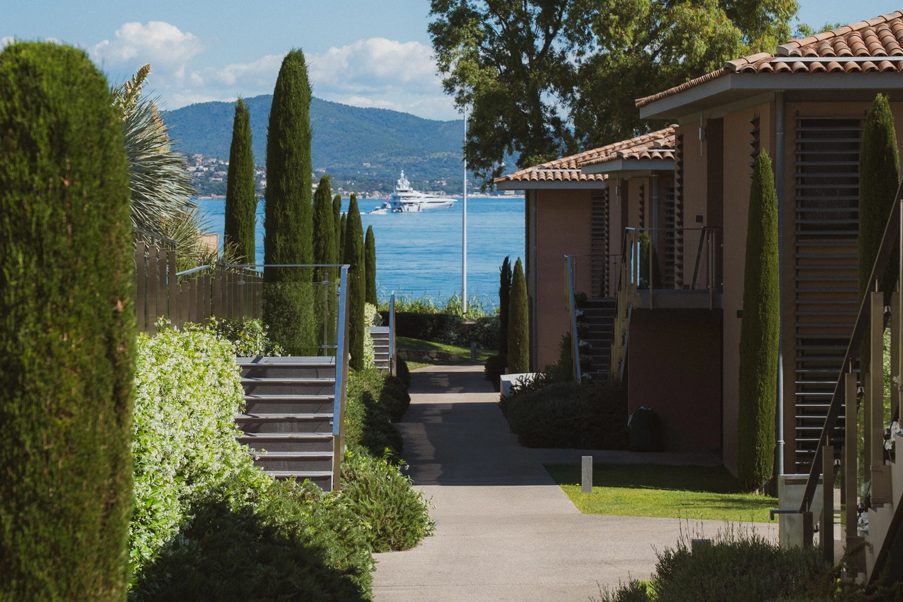 Chemin entre des maisons méditerranéennes bordé de cyprès menant à une vue sur la mer avec un yacht et des collines en arrière-plan.