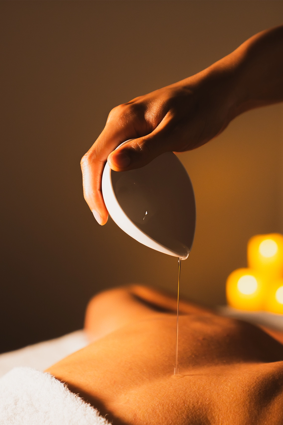Une main verse de l'huile sur le dos nu d'une personne allongée, avec des bougies allumées en arrière-plan pour une ambiance de spa.