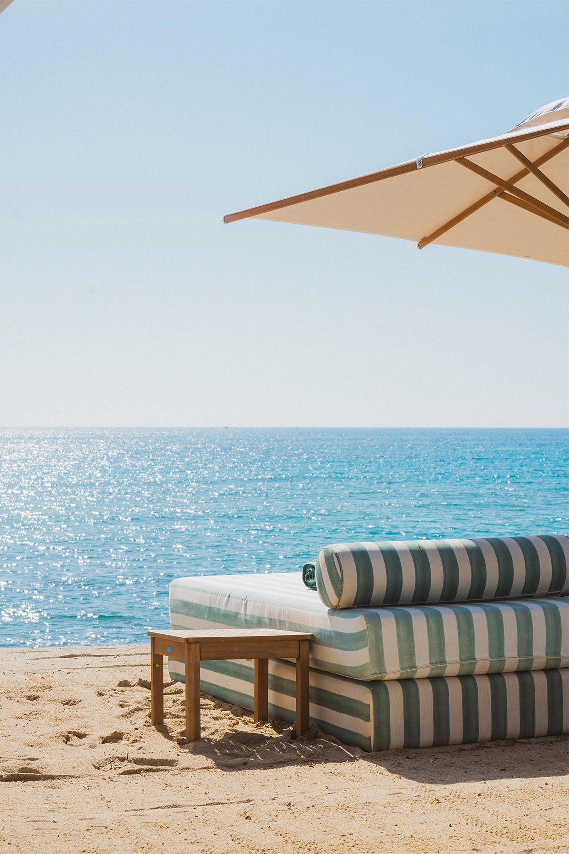 Lit de plage rayé vert et blanc avec une petite table en bois sur le sable face à la mer sous un parasol beige.