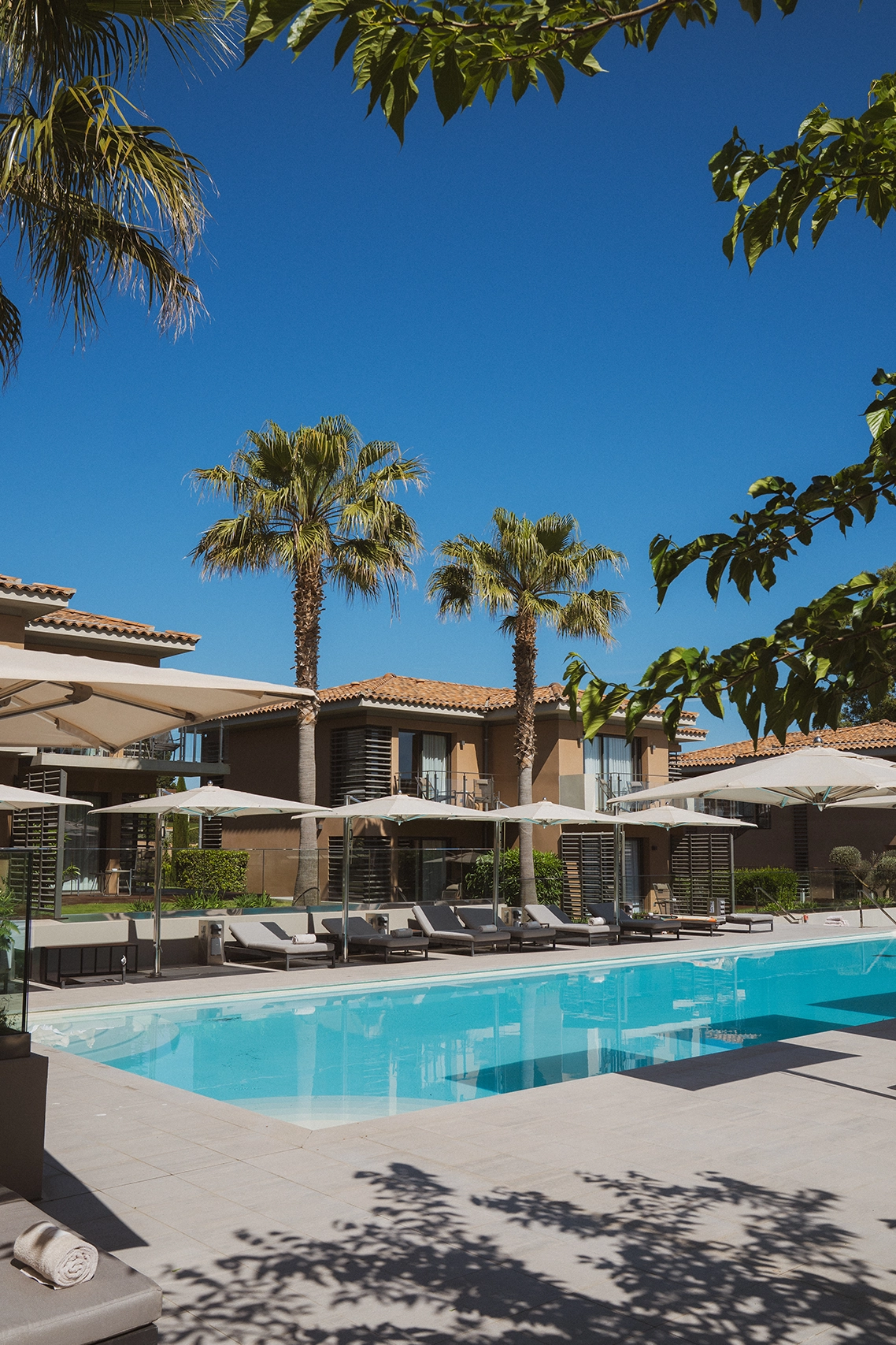Piscine extérieure avec chaises longues et parasols, entourée de palmiers et de bâtiments résidentiels sous un ciel bleu clair.