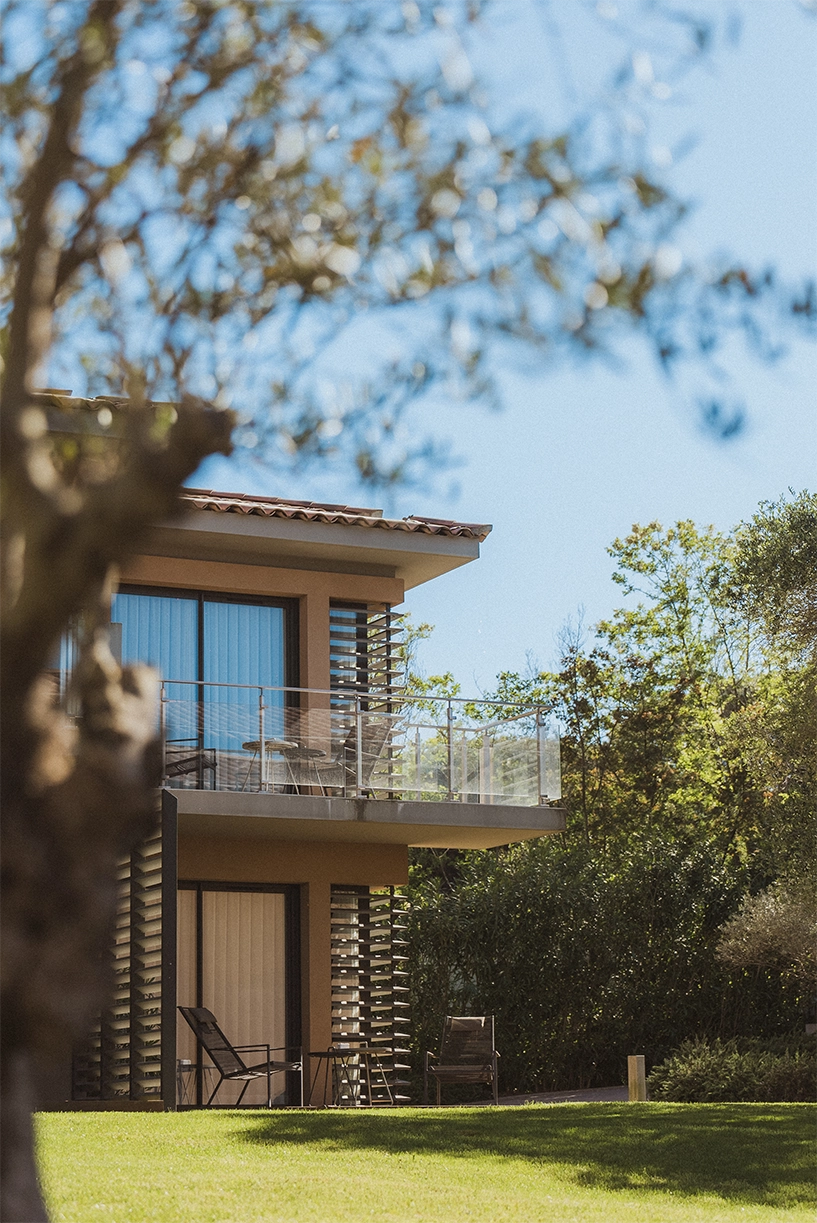 Maison moderne à deux étages avec balcon vitré, chaises et tables, entourée d'arbres et pelouse verte.