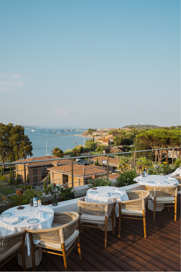 Terrasse avec tables dressées avec nappes blanches et chaises en bois, vue sur la mer avec des bateaux et des maisons en arrière-plan sous un ciel bleu.
