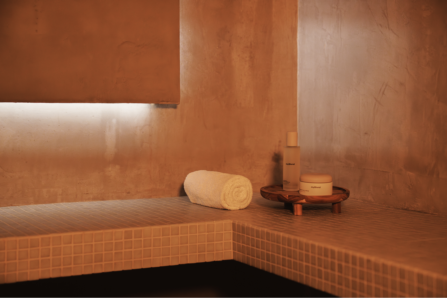 Serviette roulée blanche et deux produits de soin myBlend sur un plateau en bois dans un décor de spa aux tons chauds.