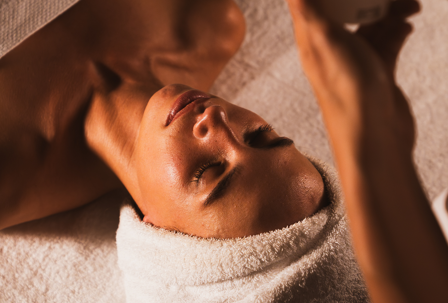Person allongée, yeux fermés, recevant un soin du visage avec une serviette blanche autour de la tête.