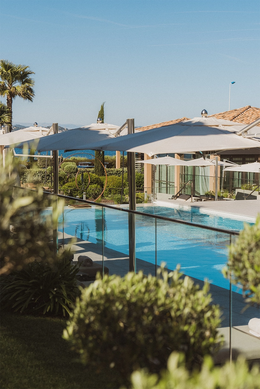 Piscine extérieure avec parasols blancs et plantes, vue vers un bâtiment aux toits en tuiles sous un ciel bleu.