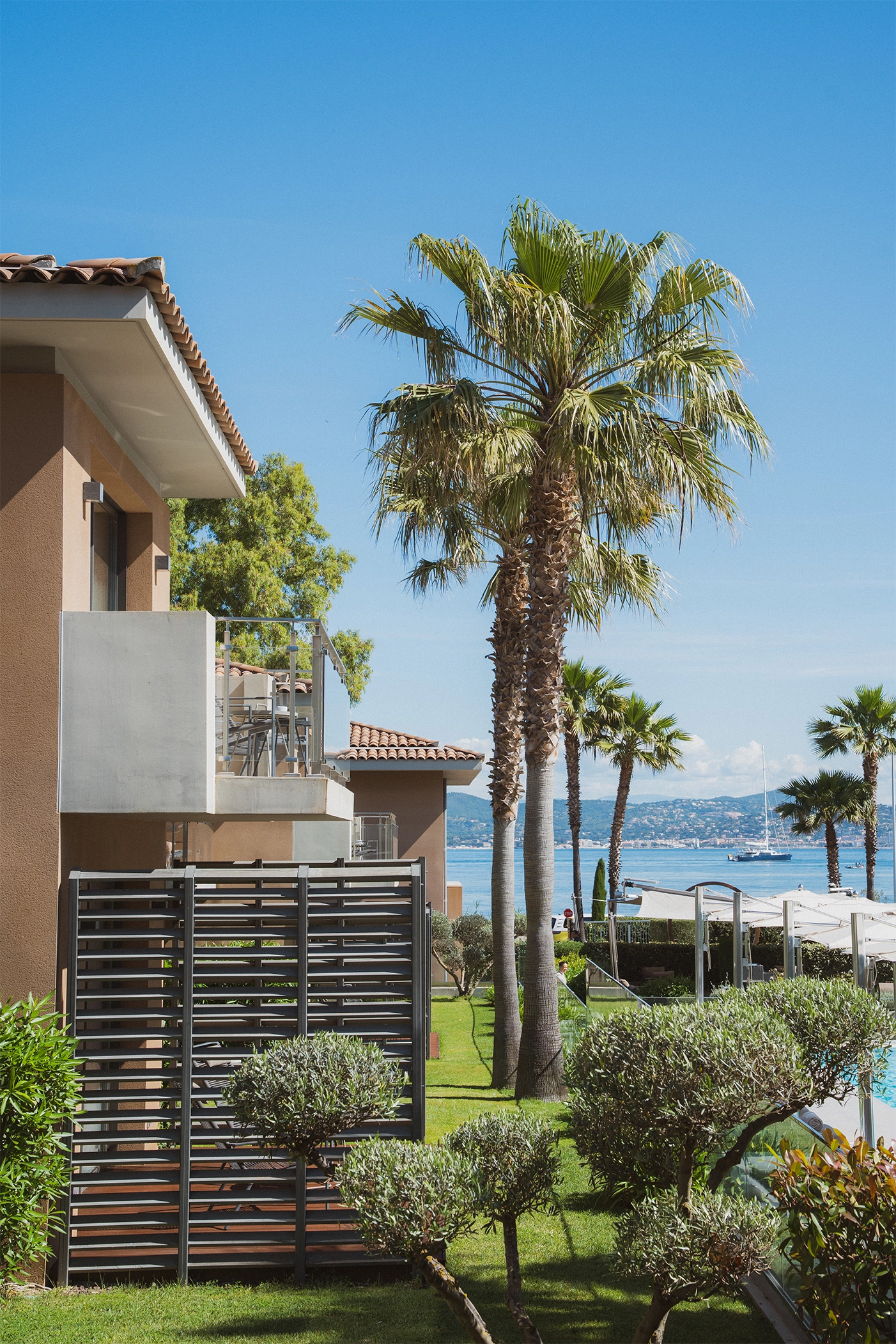 Vue sur des bâtiments modernes avec balcons, palmiers et petits arbustes dans un jardin vert, face à la mer avec un voilier et des collines à l'arrière-plan.