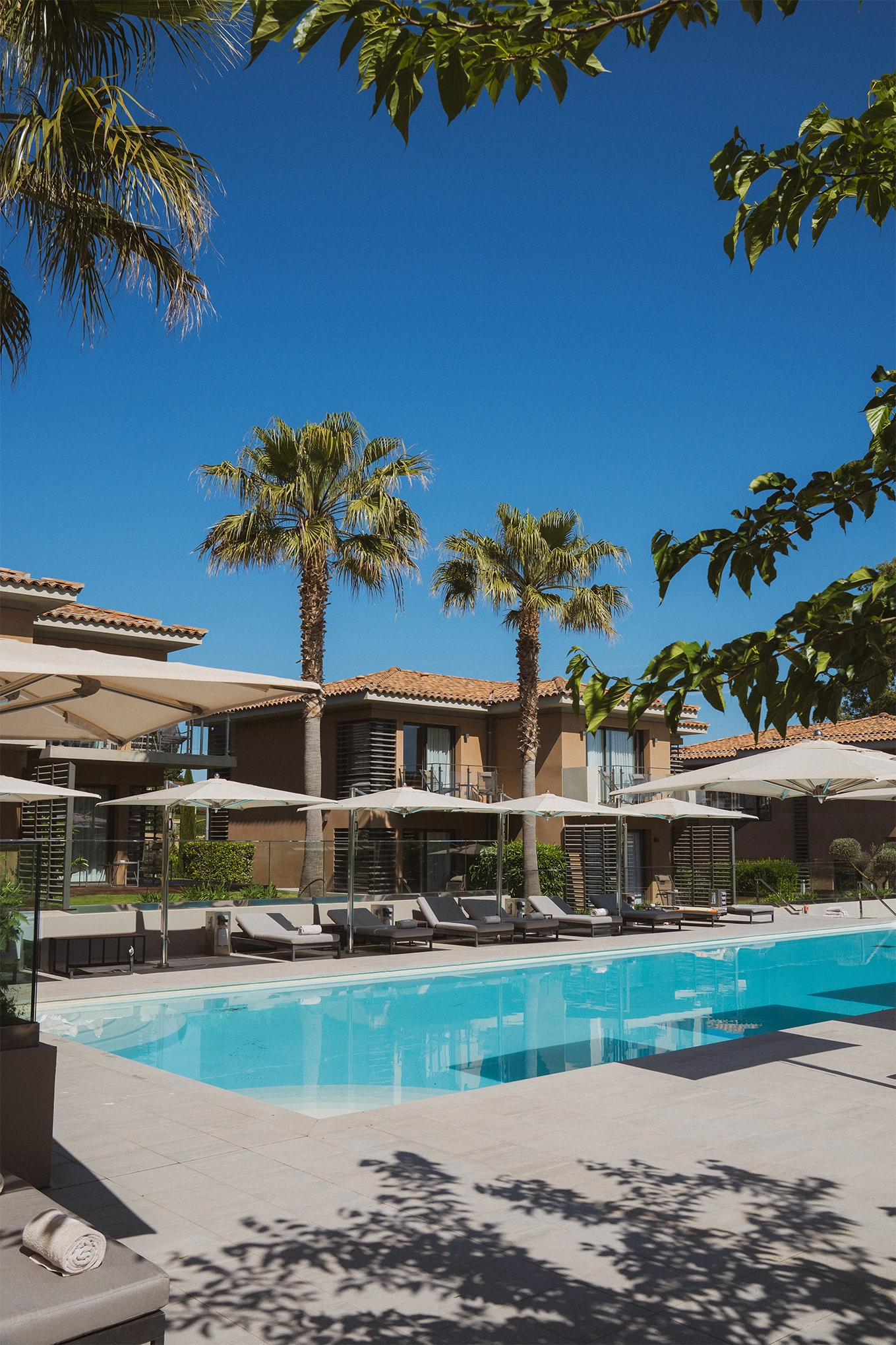 Piscine extérieure entourée de chaises longues avec parasols et palmiers devant un bâtiment de style méditerranéen sous un ciel bleu.