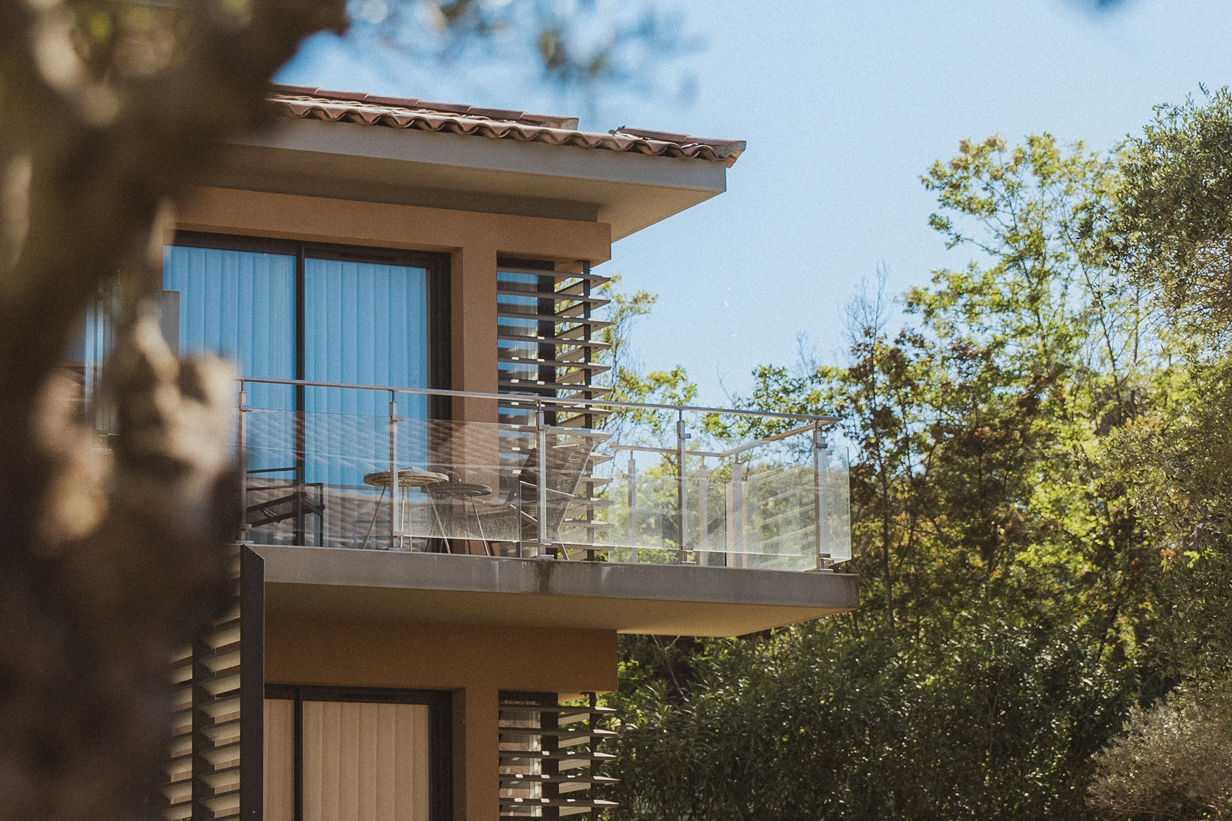Balcon d'un bâtiment moderne avec garde-corps en verre, deux tabourets et une table, entouré d'arbres verts sous un ciel bleu clair.