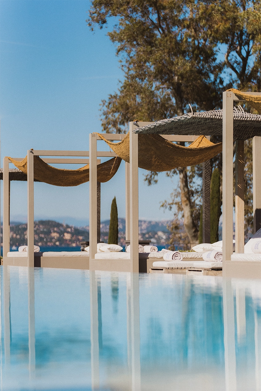 Piscine à débordement avec lits de soleil sous des pergolas et vue sur des collines et des arbres par une journée ensoleillée.