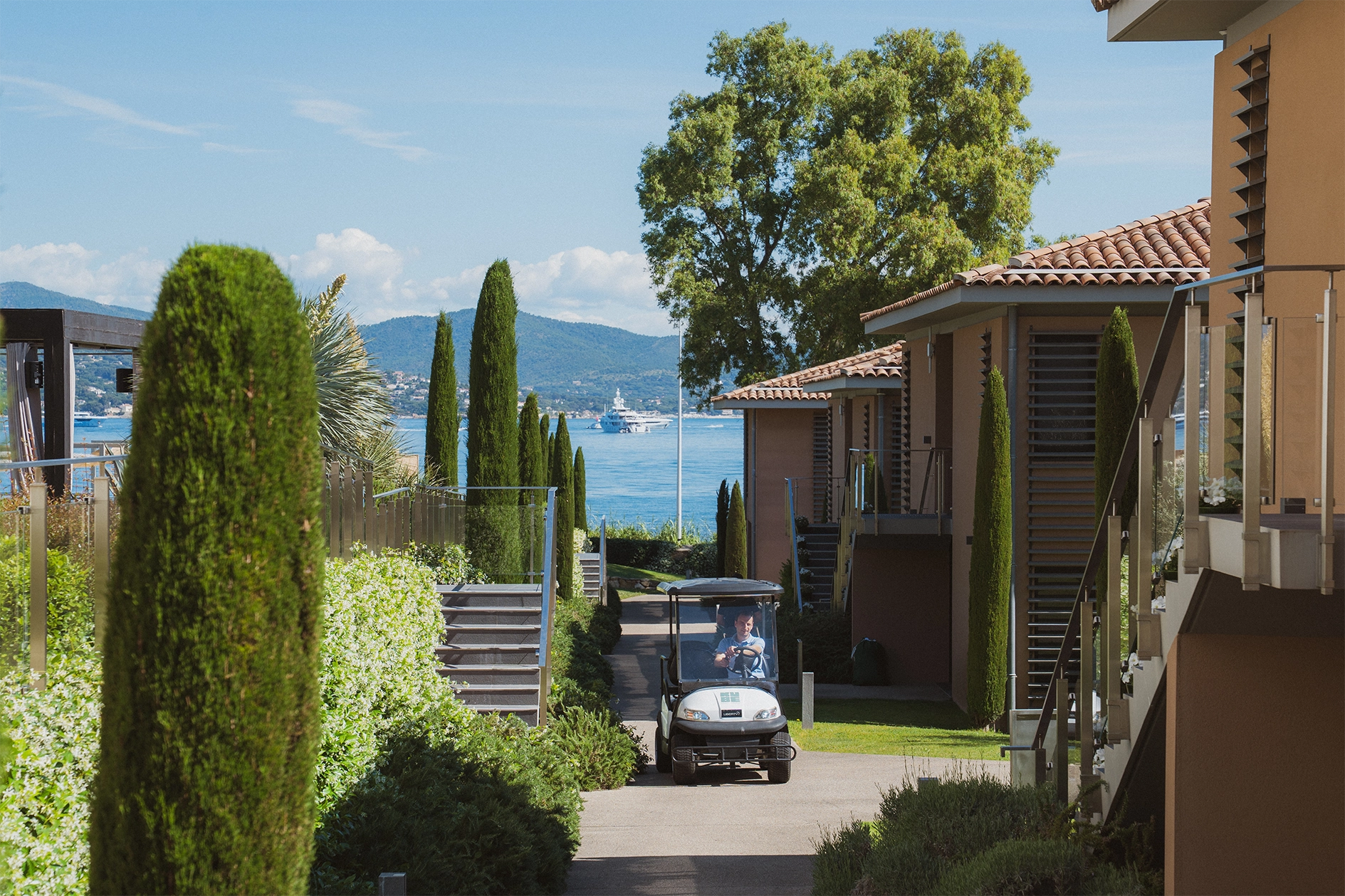 Petit véhicule électrique circulant sur une allée bordée de cyprès entre des bâtiments résidentiels avec vue sur la mer et les montagnes à l'arrière.