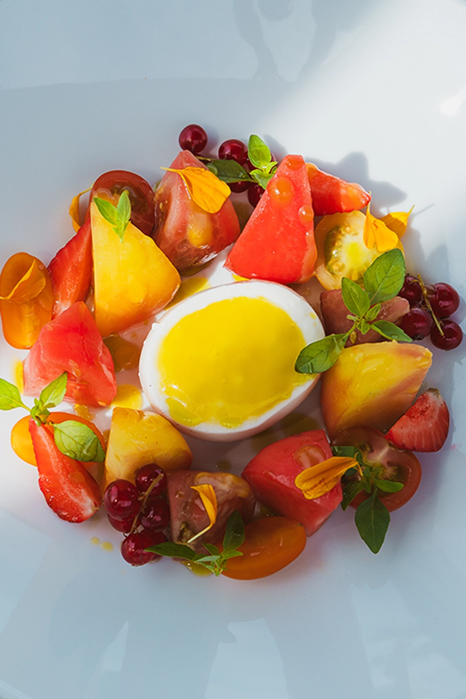 Oeuf mollet avec sauce jaune entouré de morceaux de tomates multicolores, fraises, groseilles, feuilles vertes et pétales jaunes dans une assiette blanche.