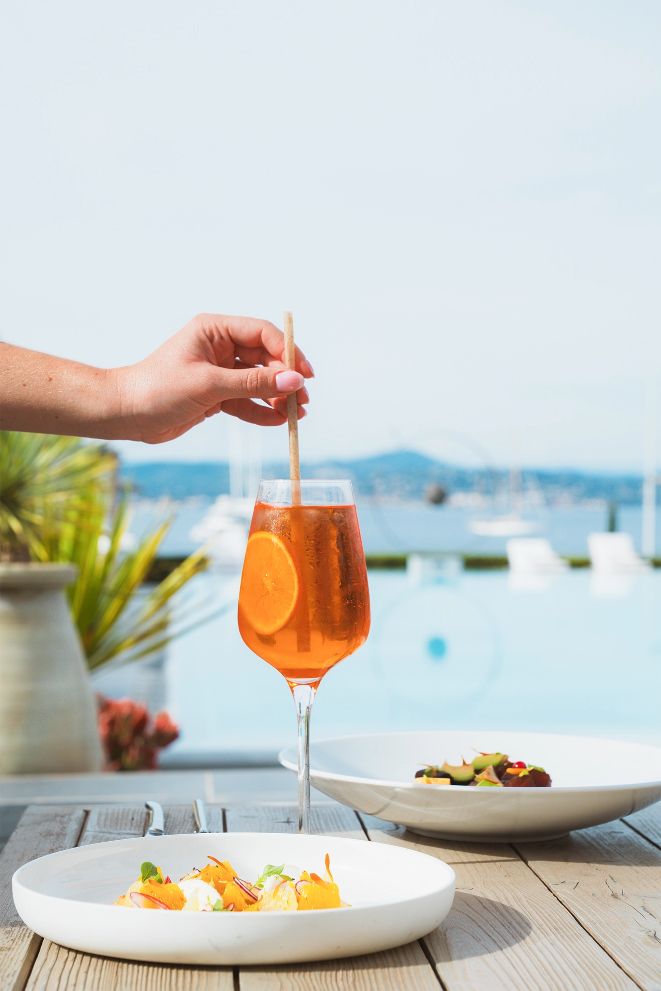 Une main féminine tient un verre de cocktail orange avec une tranche d'orange au bord d'une table en bois avec deux assiettes et vue sur une piscine et la mer.