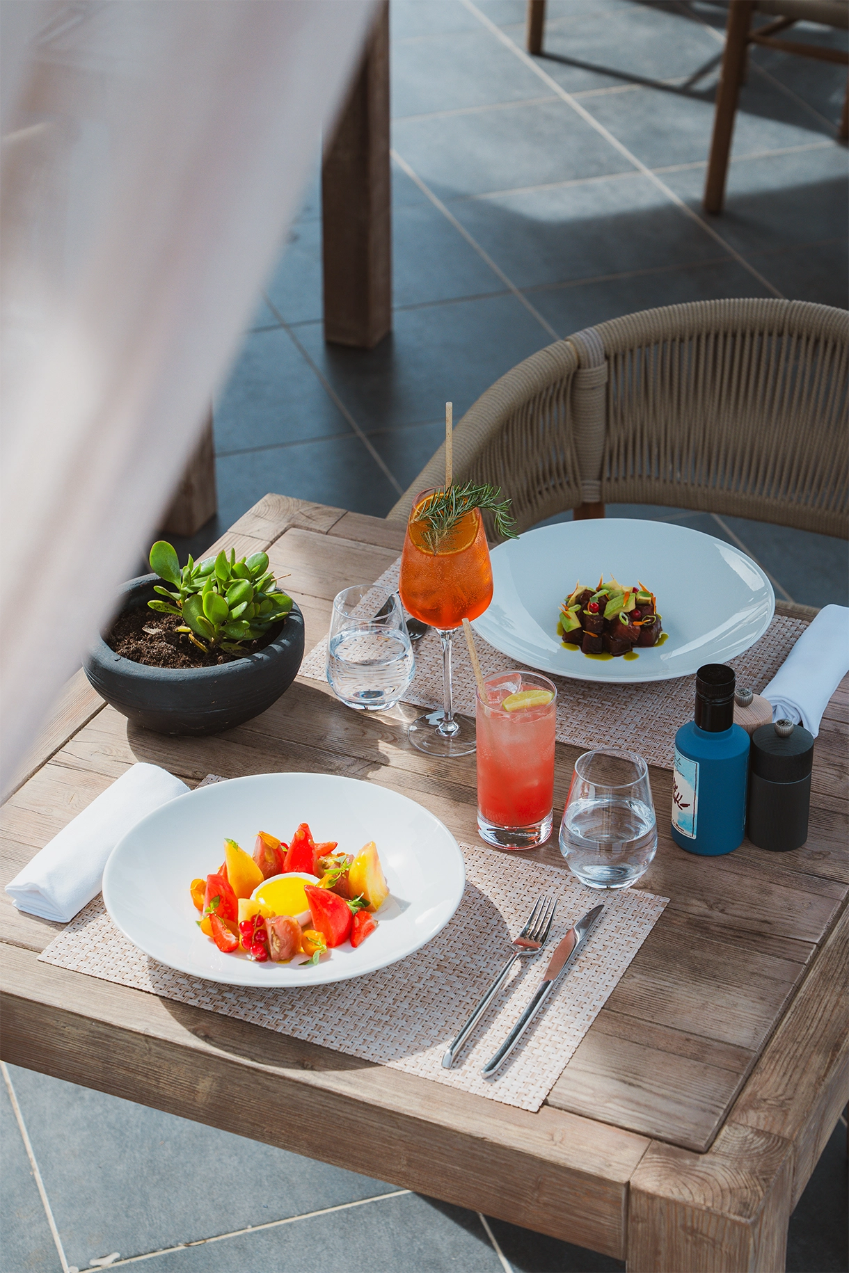 Table en bois avec deux assiettes de plats gastronomiques, deux verres d'eau, deux cocktails colorés et une plante verte en pot.