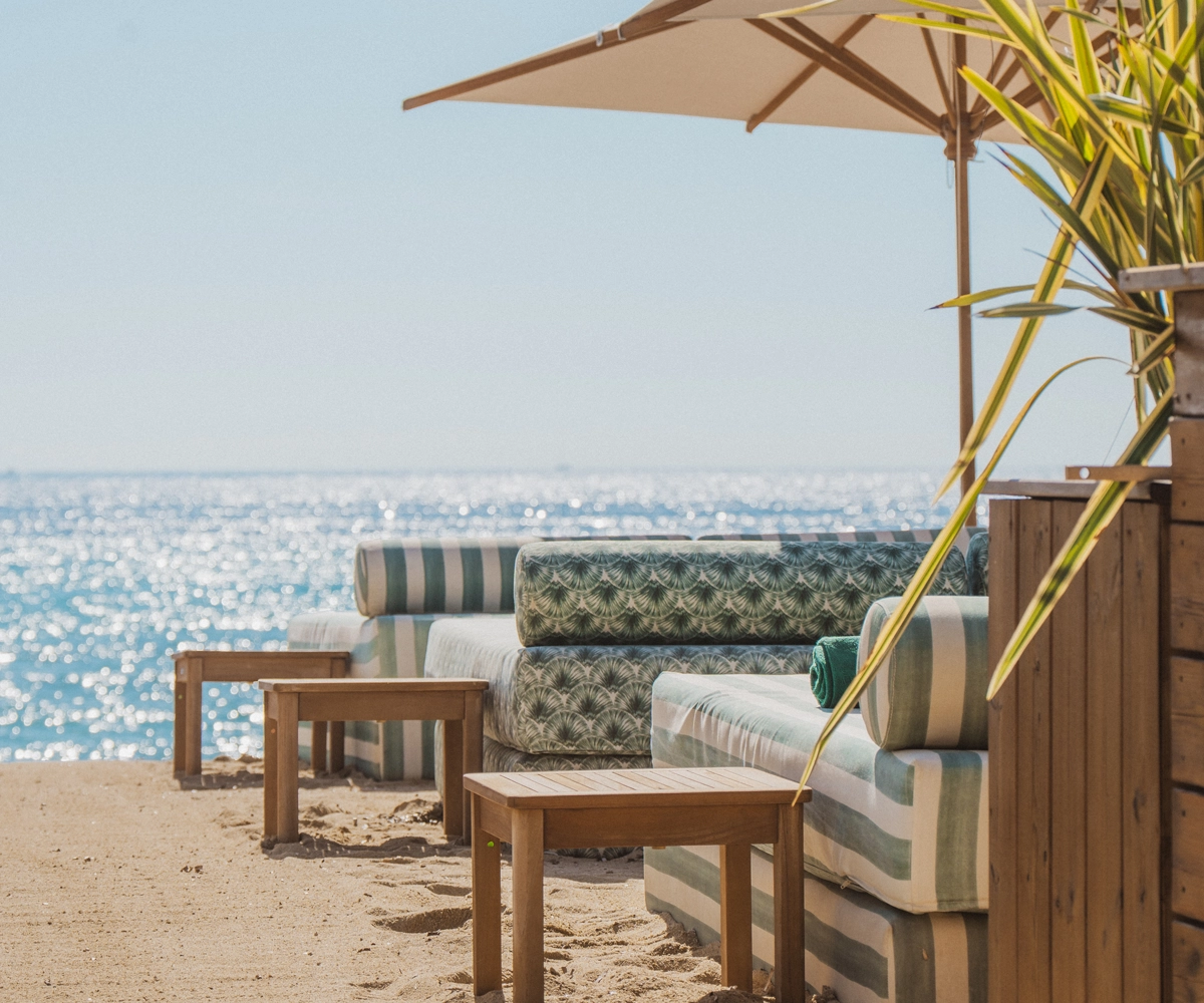 Canapés et tables basses en bois installés sur une plage avec vue sur la mer scintillante et un parasol beige.