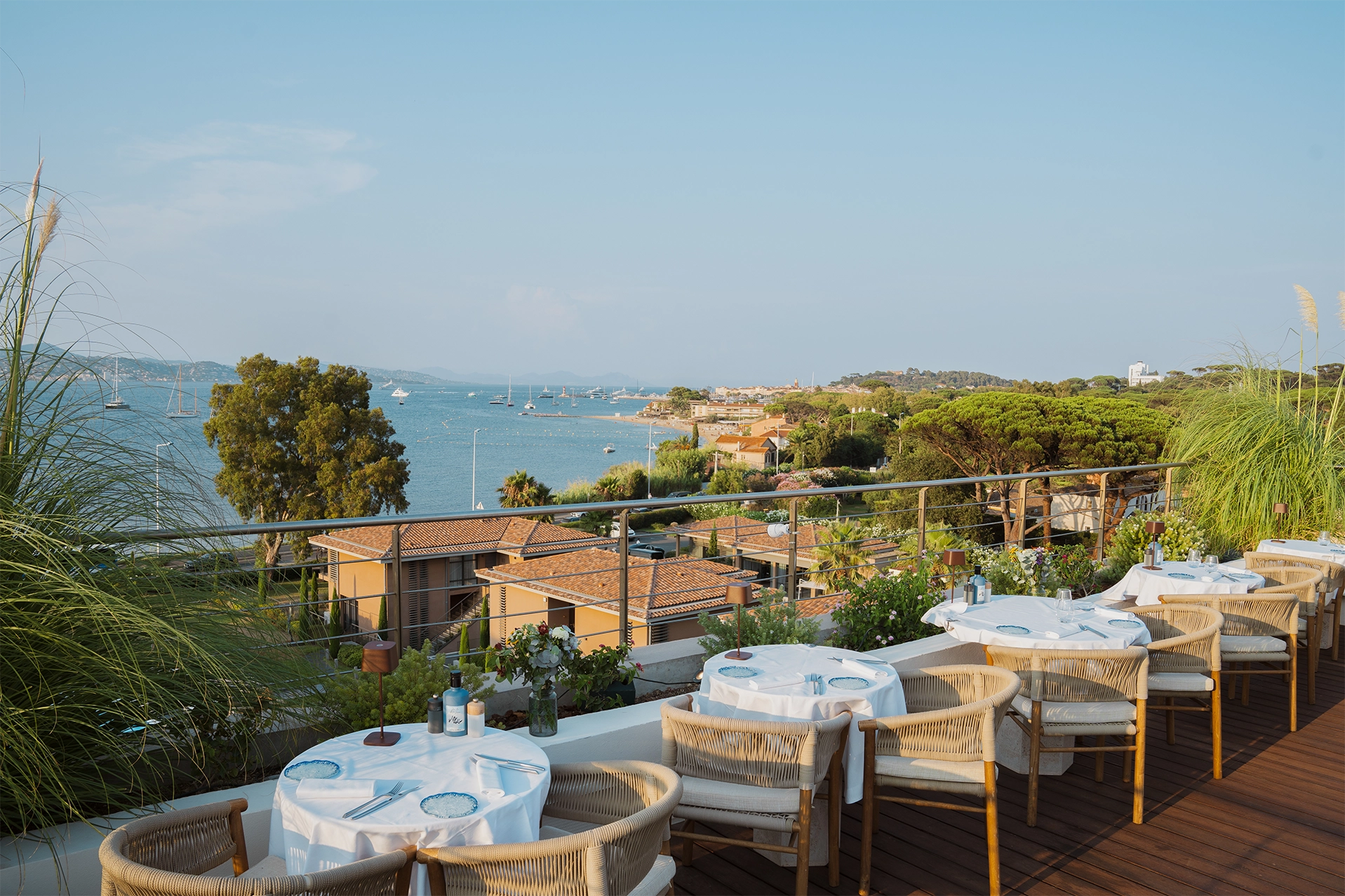 Terrasse de restaurant avec tables dressées surplombant la mer et des bateaux ancrés près de la côte sous un ciel bleu clair.