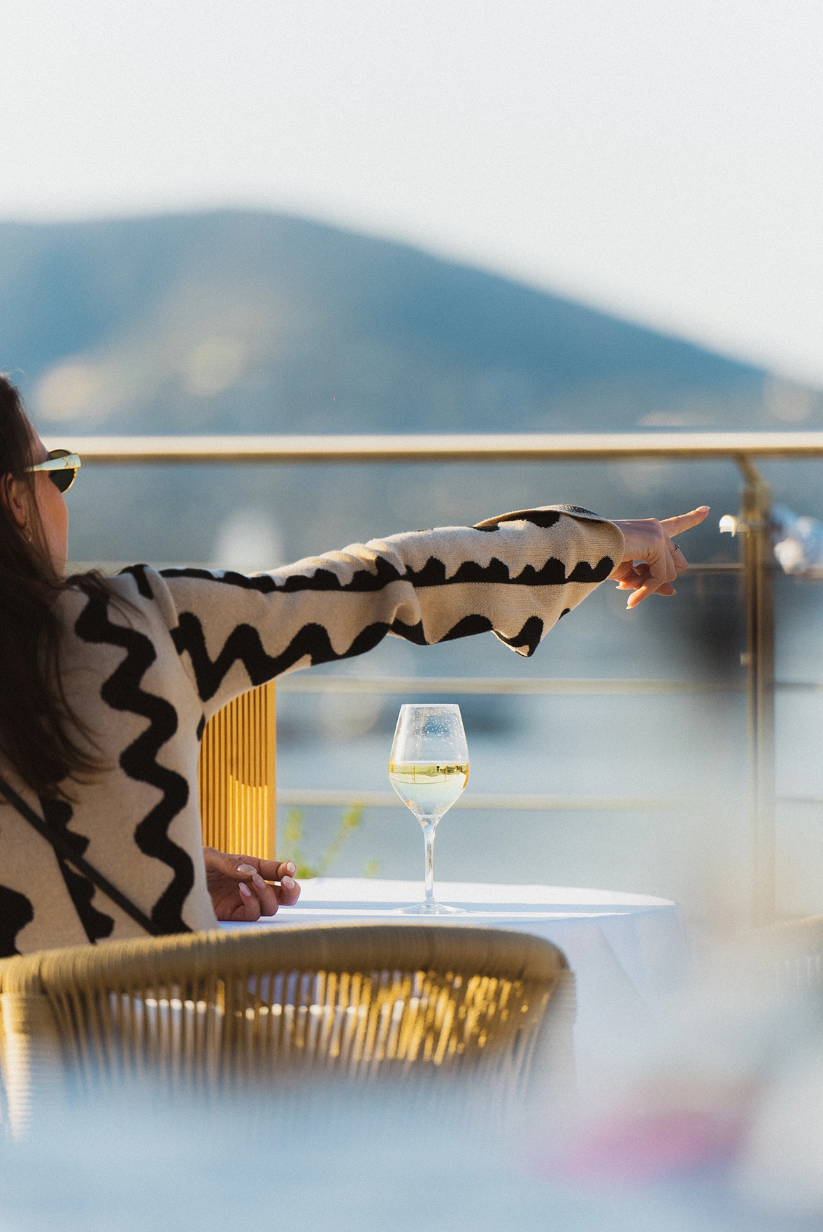 Person assise à une table en extérieur avec un verre de vin blanc, pointant du doigt vers l'horizon avec une montagne floue en arrière-plan.