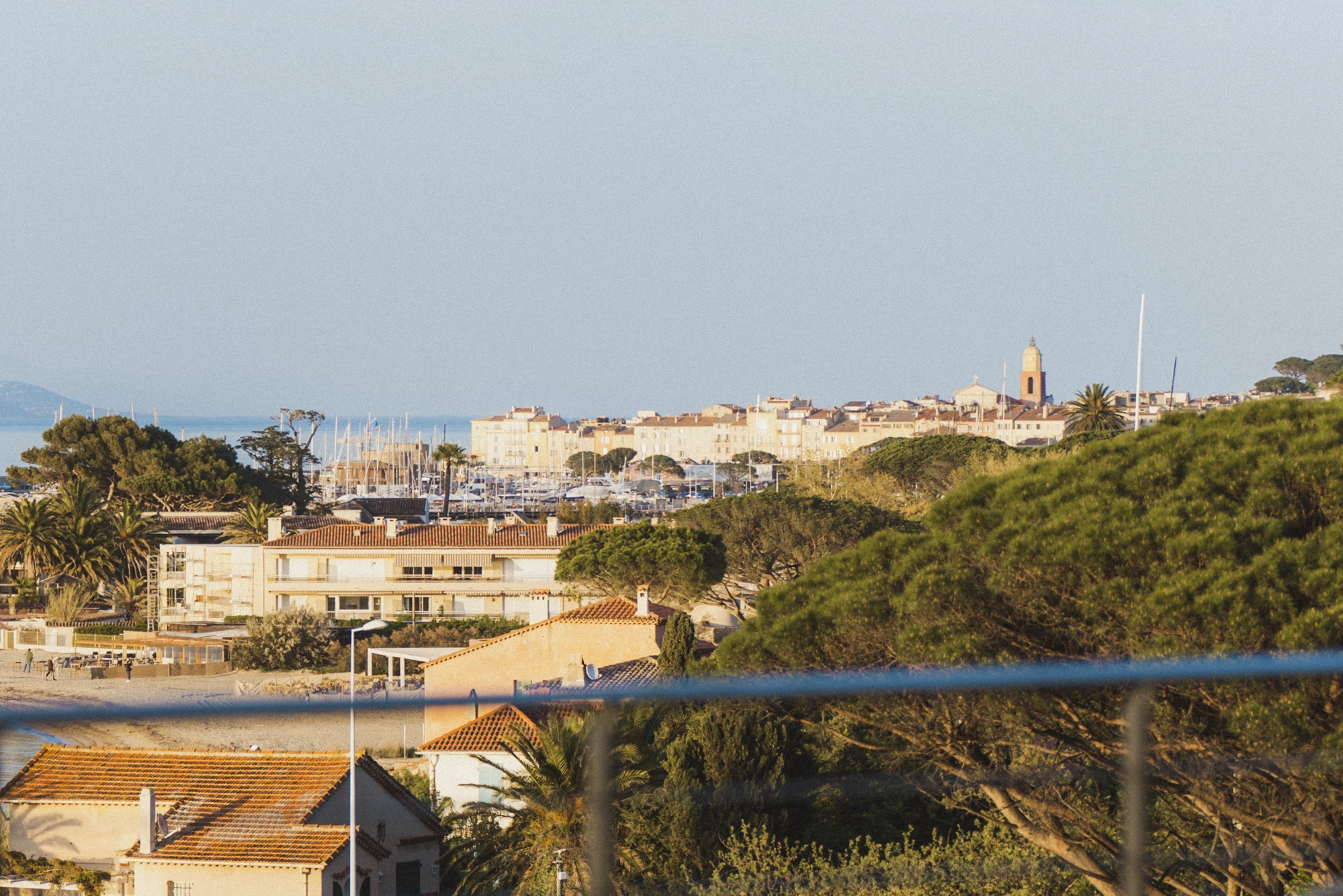 Vue panoramique de Saint-Tropez avec des toits en tuiles, des arbres verts, un port de plaisance et la mer en arrière-plan au coucher du soleil.