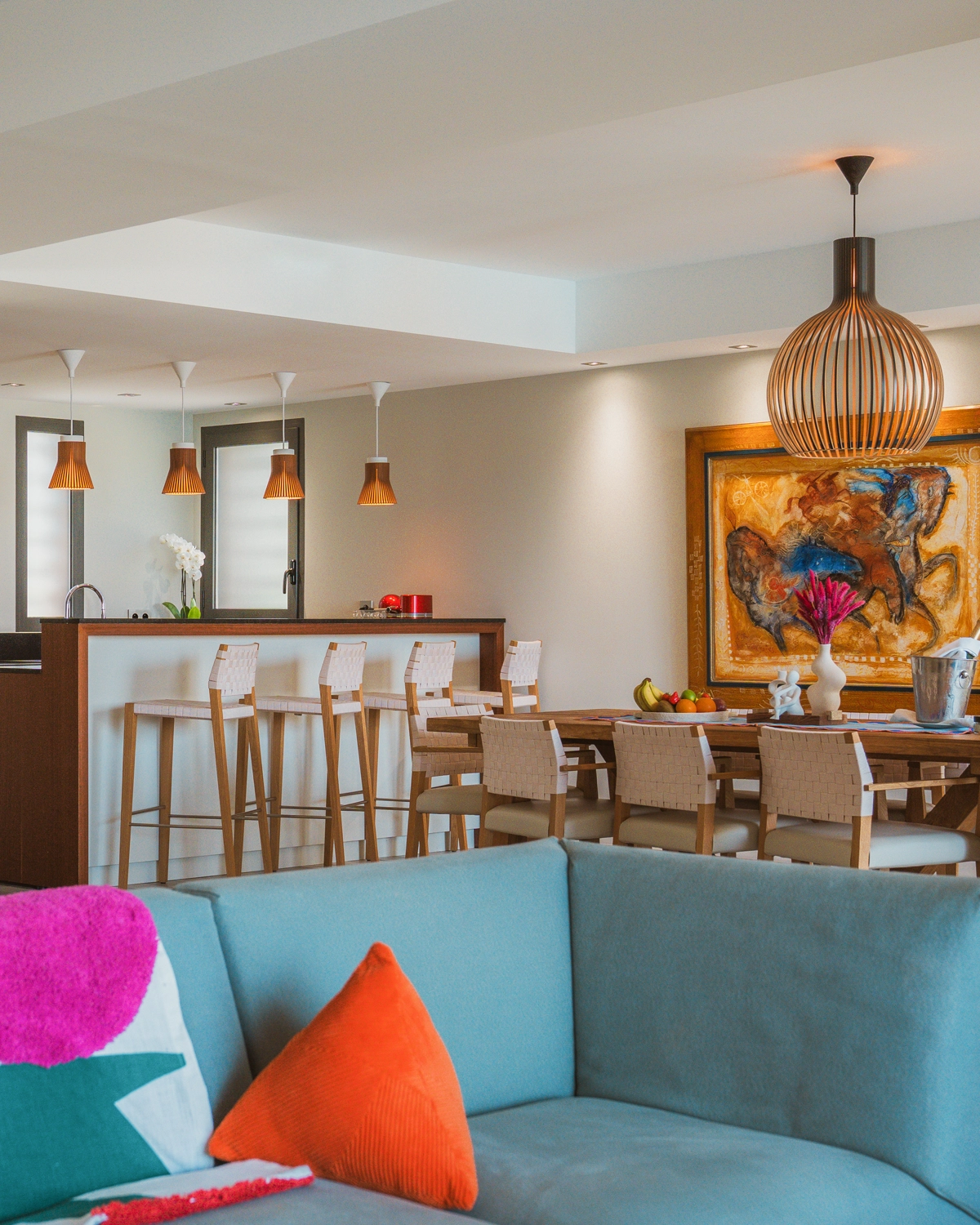 Salon moderne avec canapé bleu, coussins colorés, table à manger en bois avec chaises tressées, bar avec tabourets et suspensions en bois.