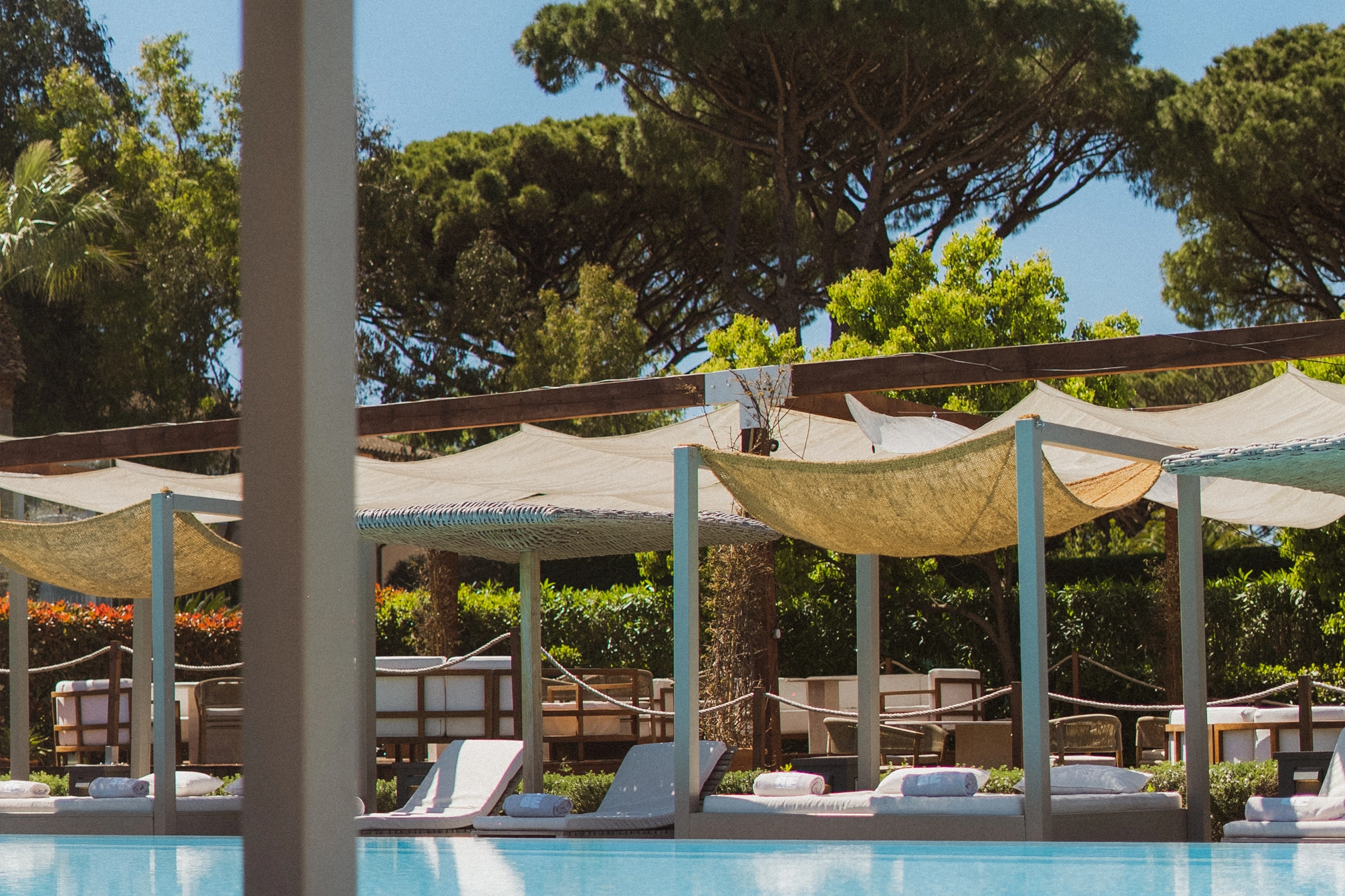 Piscine avec transats et cannisses fournissant de l’ombre, entourée de verdure et de grands arbres sous un ciel bleu clair.