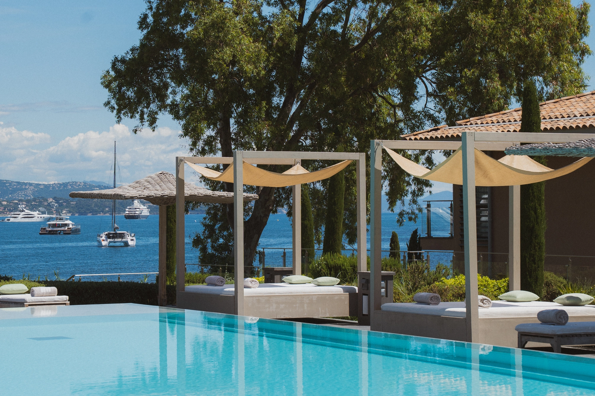 Piscine avec lits de jour ombragés et vue sur la mer avec plusieurs bateaux au loin.