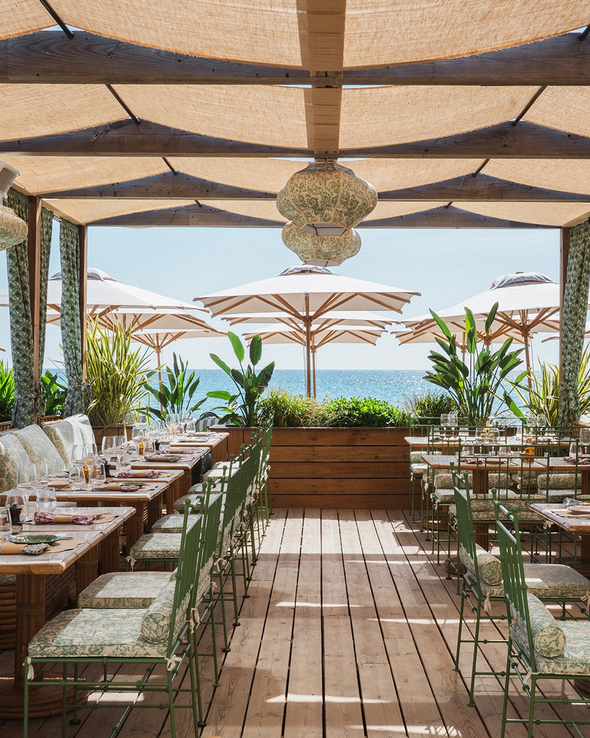 Terrasse de restaurant en bord de mer avec tables en bois, chaises vertes à coussin et parasols blancs sous un auvent ombragé.