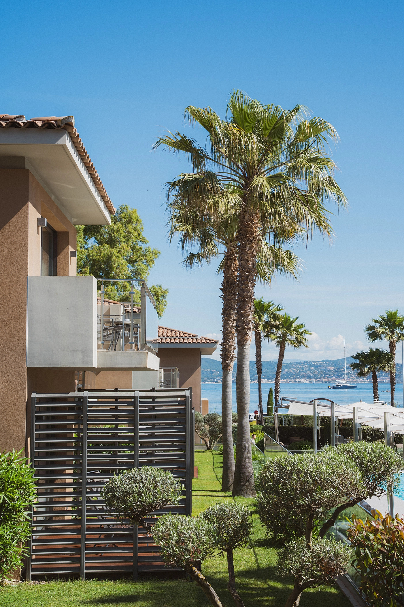 Vue sur un jardin avec palmiers et buissons devant des bâtiments modernes, avec la mer et un voilier en arrière-plan sous un ciel bleu clair.