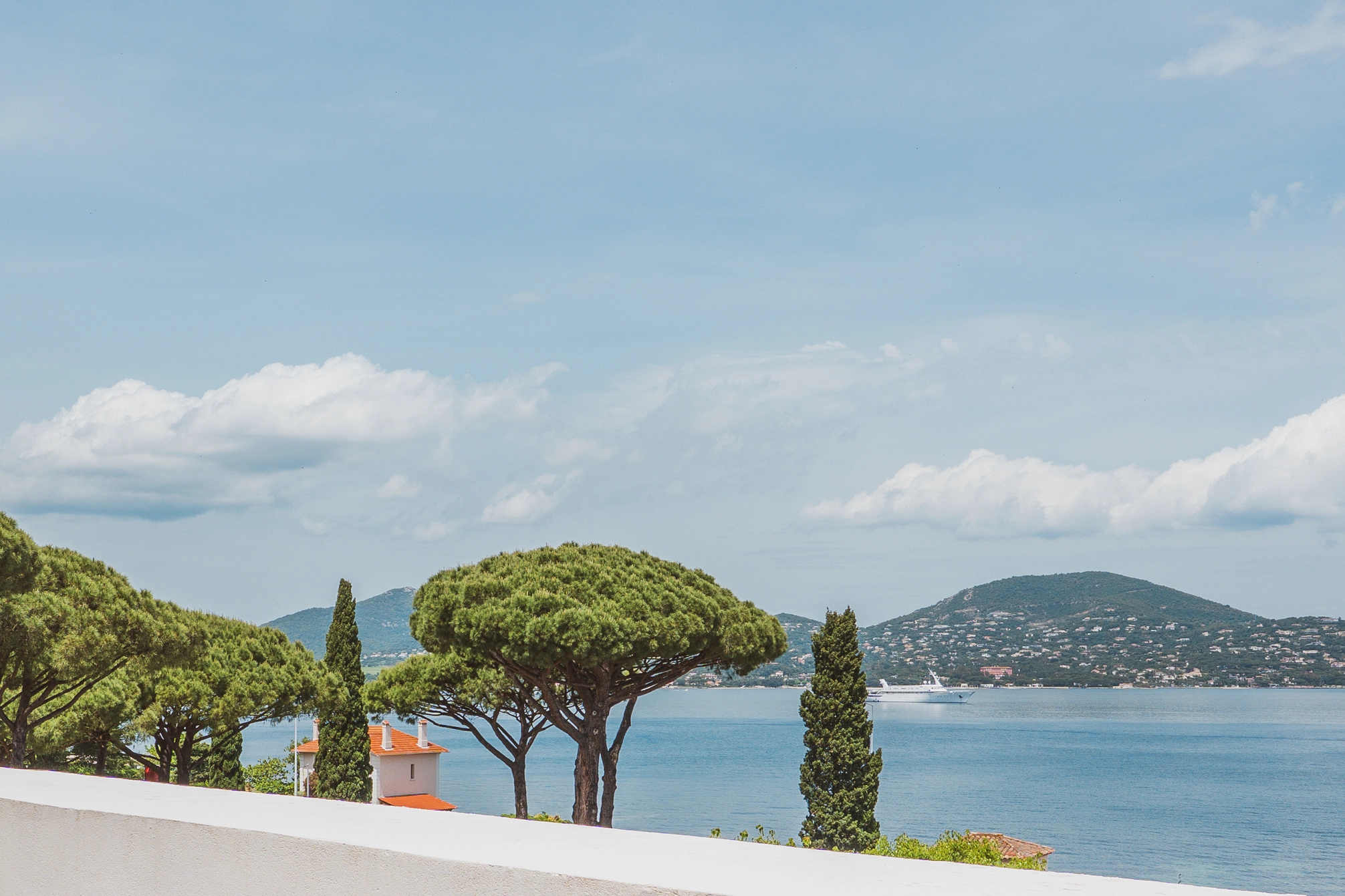 Vue dégagée sur un paysage côtier avec des pins méditerranéens, une maison au toit rouge, une mer calme et un yacht blanc à l'horizon devant des collines.