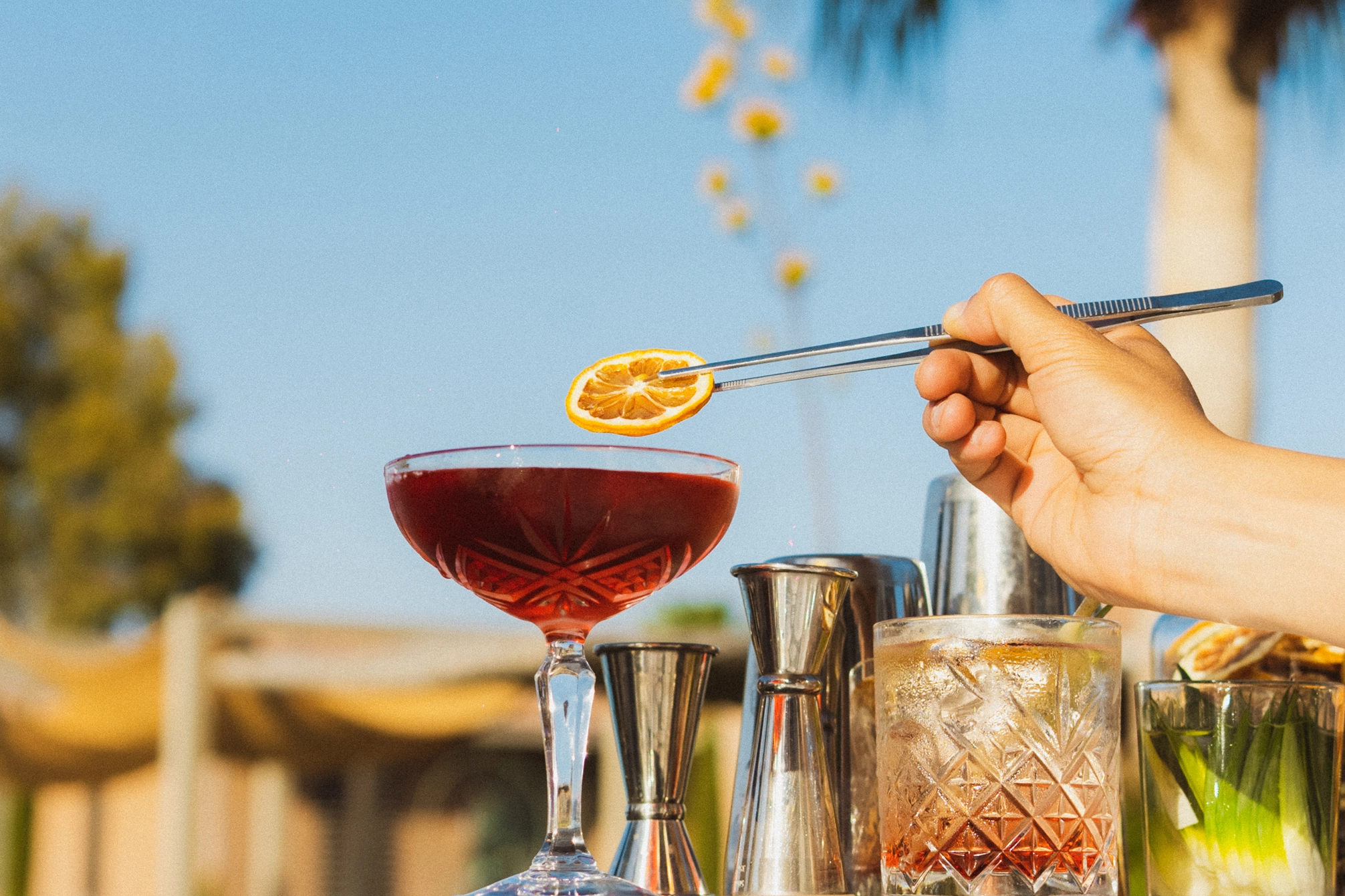 Une main ajoute une tranche d'orange séchée avec une pince à un cocktail rouge dans un verre à pied, avec des outils de bar en arrière-plan.