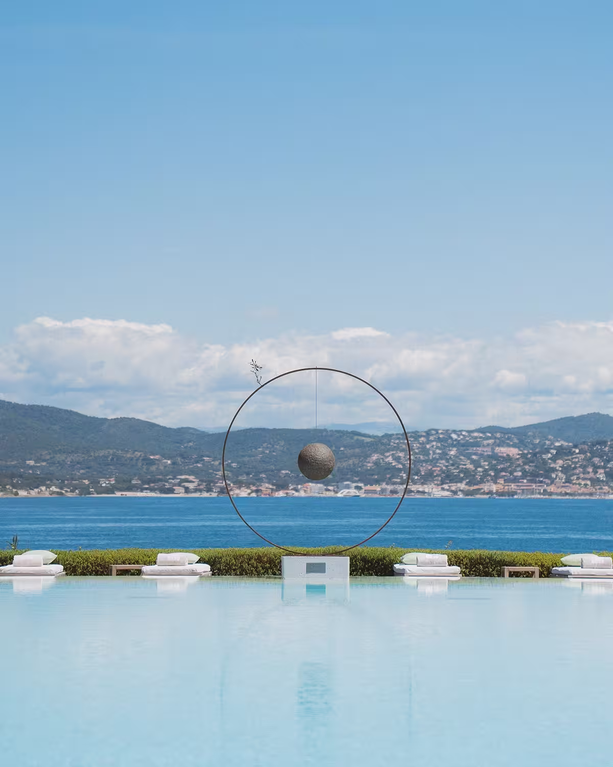 Piscine à débordement avec une sculpture circulaire et une sphère suspendue, vue sur la mer et les collines à l'arrière-plan.