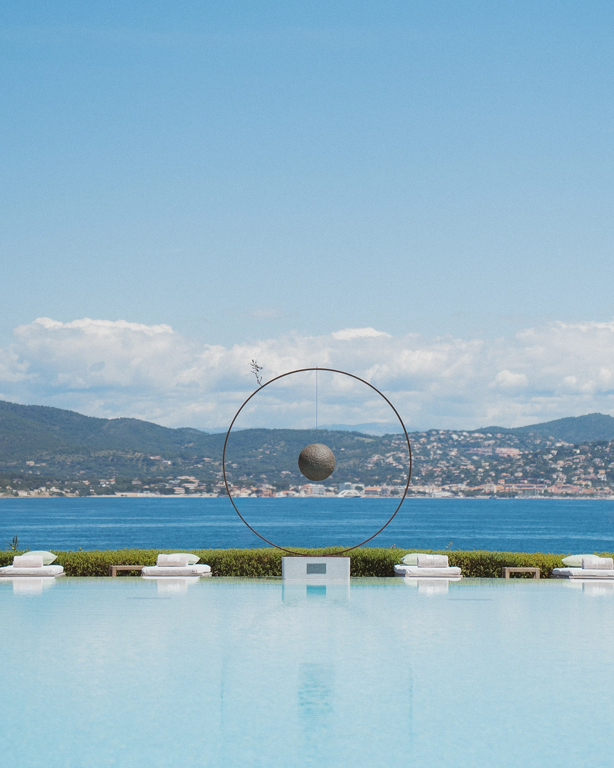 Piscine à débordement avec une sculpture circulaire et une sphère suspendue, vue sur la mer et les collines à l'arrière-plan.