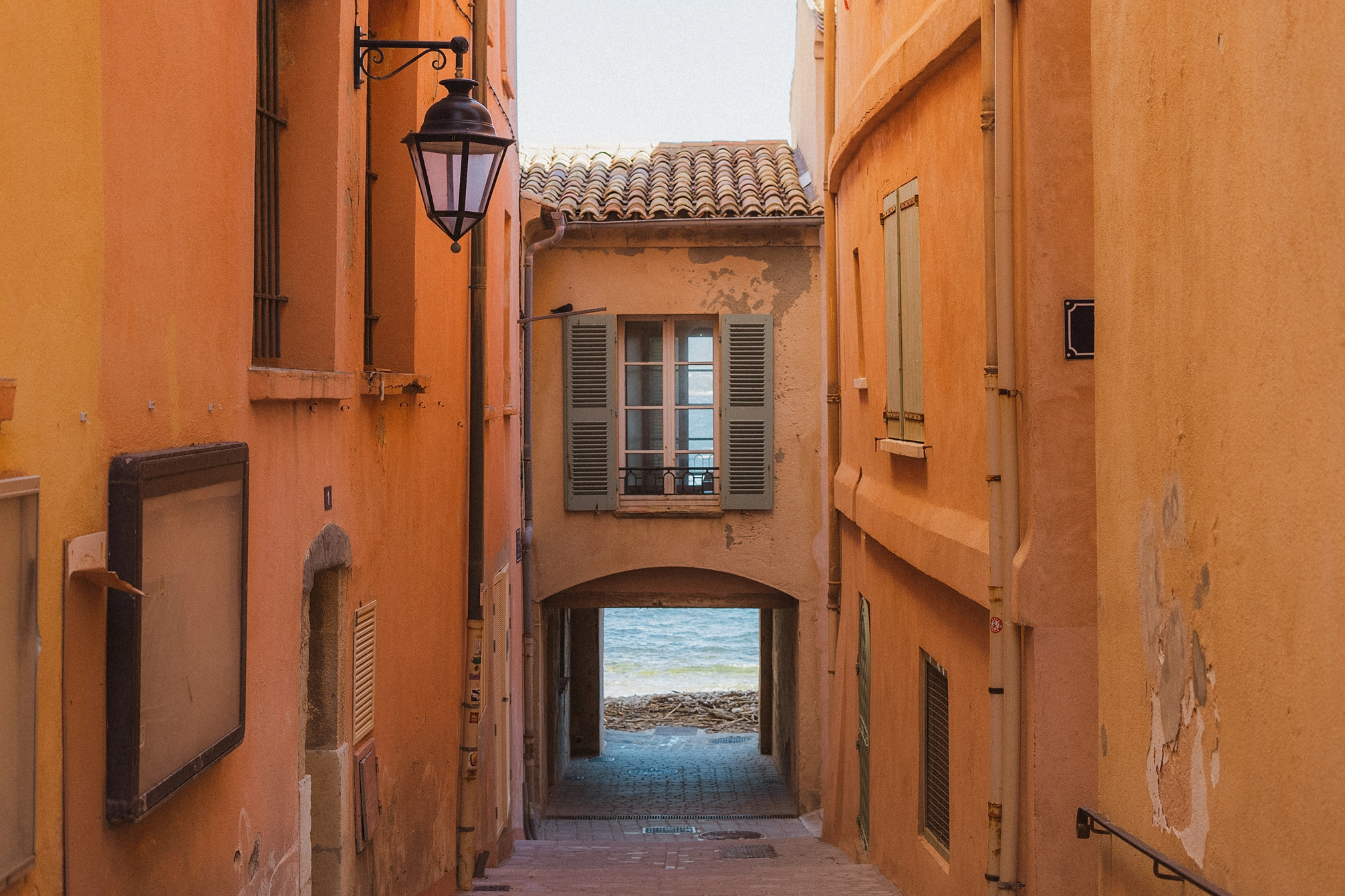 Ruelle étroite aux murs orange menant à une arche avec vue sur la mer et une fenêtre à volets gris clair.